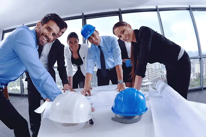 A group of professionals in hard hats and business attire collaborates around a table covered with blueprints, showcasing teamwork in a modern office setting.