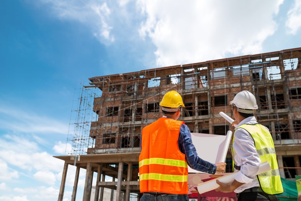 Two construction workers, one in an orange vest and yellow hard hat and the other in a white hard hat, review blueprints in front of a building under construction against a clear blue sky.