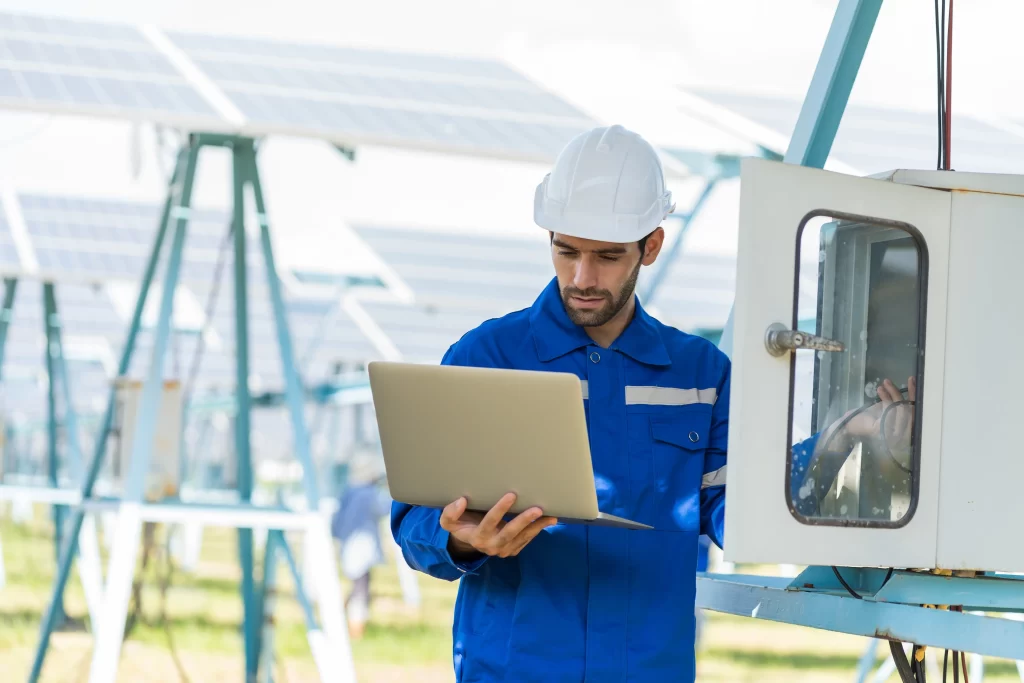 A technician in a blue uniform and hard hat is using a laptop while inspecting equipment near solar panels. The setting is an outdoor renewable energy facility.