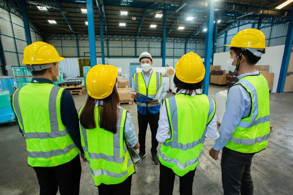 A group of five individuals in safety vests and hard hats listens attentively to a speaker in a warehouse setting. The speaker is wearing a mask, indicating a focus on safety protocols.