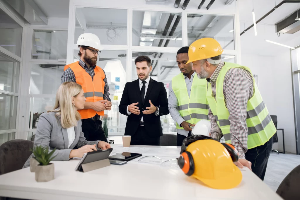 A group of professionals in safety gear discusses a project around a table, with a woman using a tablet and men in hard hats and vests actively engaging in the conversation. The setting appears to be a modern office environment.