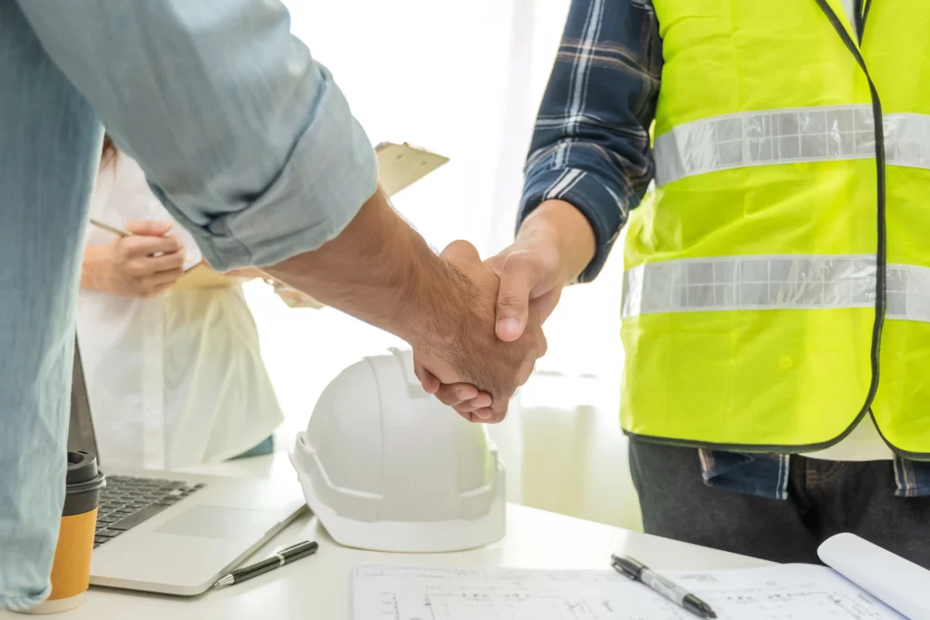 A handshake between two individuals, one wearing a safety vest, signifies a professional agreement or collaboration in a construction or engineering context. A hard hat and documents are visible on the table.