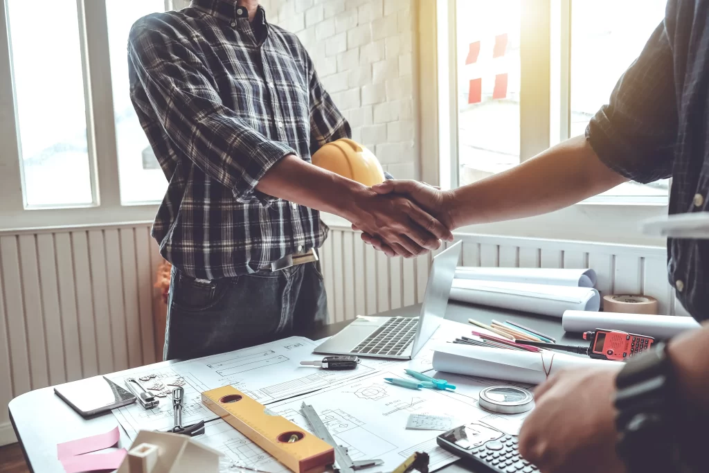 Two individuals shake hands over a table filled with architectural plans and tools, symbolizing a business agreement or collaboration.