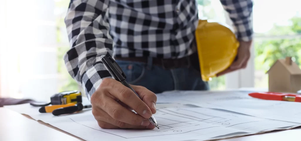A person in a checkered shirt is writing on blueprints while holding a yellow hard hat. Tools are visible on the table, indicating a construction or planning context.