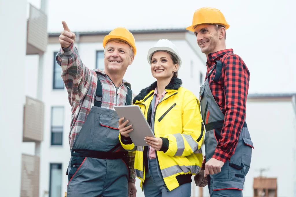 Three construction workers, one pointing and smiling, stand together outdoors, discussing plans while holding a tablet. They are wearing safety helmets and work attire.