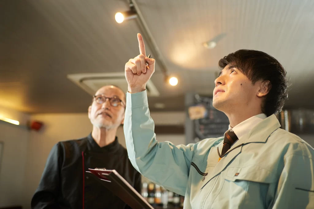 A young man in a blue work uniform points upwards while discussing something with an older man who looks on thoughtfully. The setting appears to be an indoor space, possibly a restaurant or café.