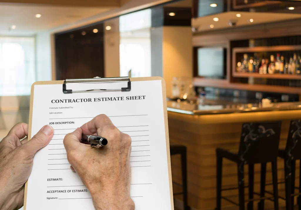 A person is writing on a contractor estimate sheet while standing in a modern kitchen with a bar area in the background. The setting suggests a home renovation or construction project.