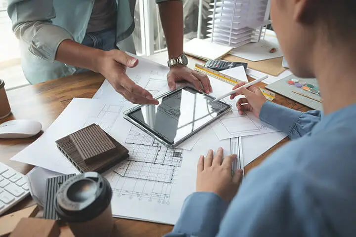 Two people are engaged in a discussion over a tablet while reviewing architectural plans and documents on a desk. A coffee cup and a stack of papers are also visible.