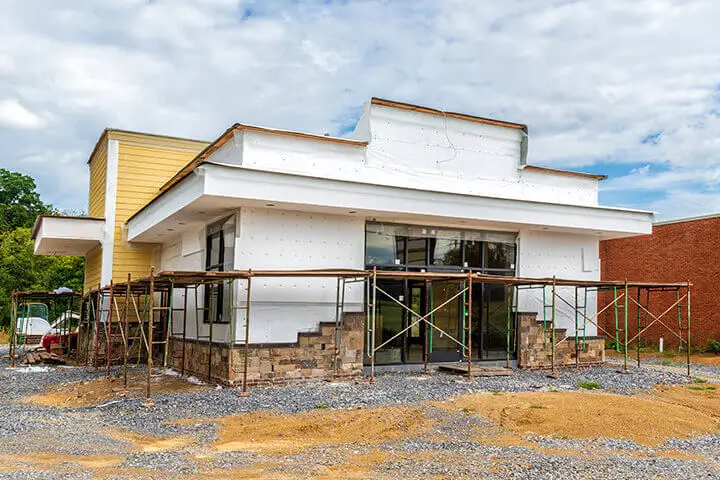 A newly constructed building with scaffolding, featuring a mix of white and yellow exterior finishes. The surrounding area is still under development, with gravel and dirt visible.