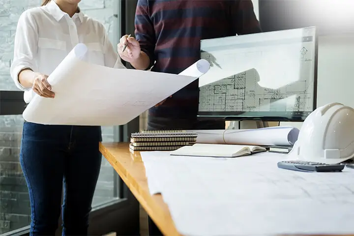 Two people discuss architectural plans while standing next to a computer and a stack of notebooks on a table. A construction helmet is also visible on the table.